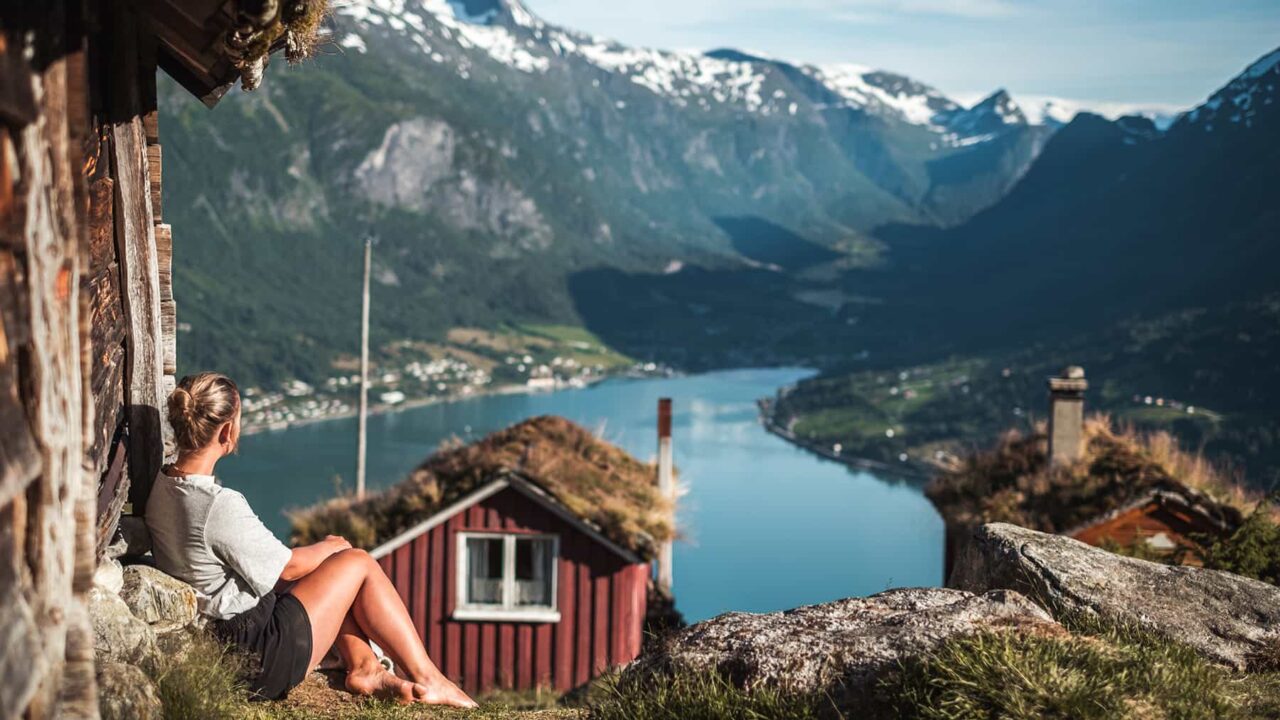 barfota kvinna som sitter i naturmiljö och blickar ut över berg, hav och en röd stuga
