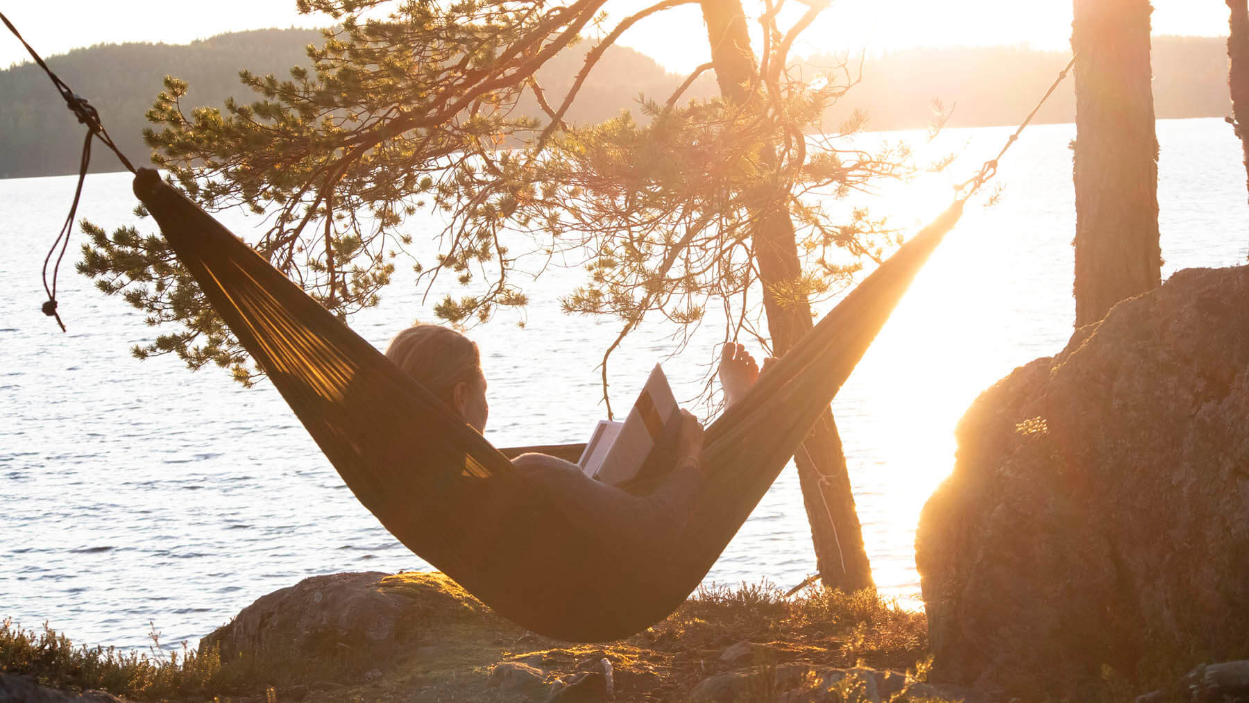 En kvinna läser bok i en hängmatta i solnedgången invid vatten.