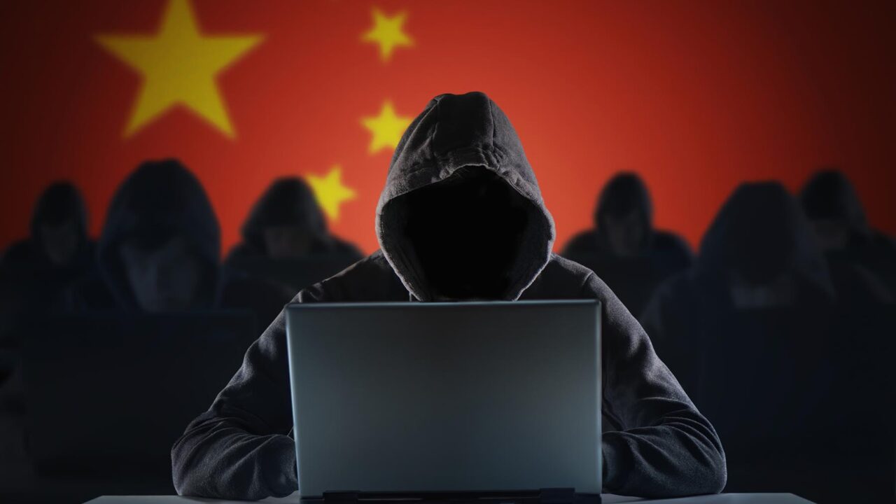 Cyberbrottsling i huva framför en laptop, i bakgrunden många fler och en stor kinesisk flagga.