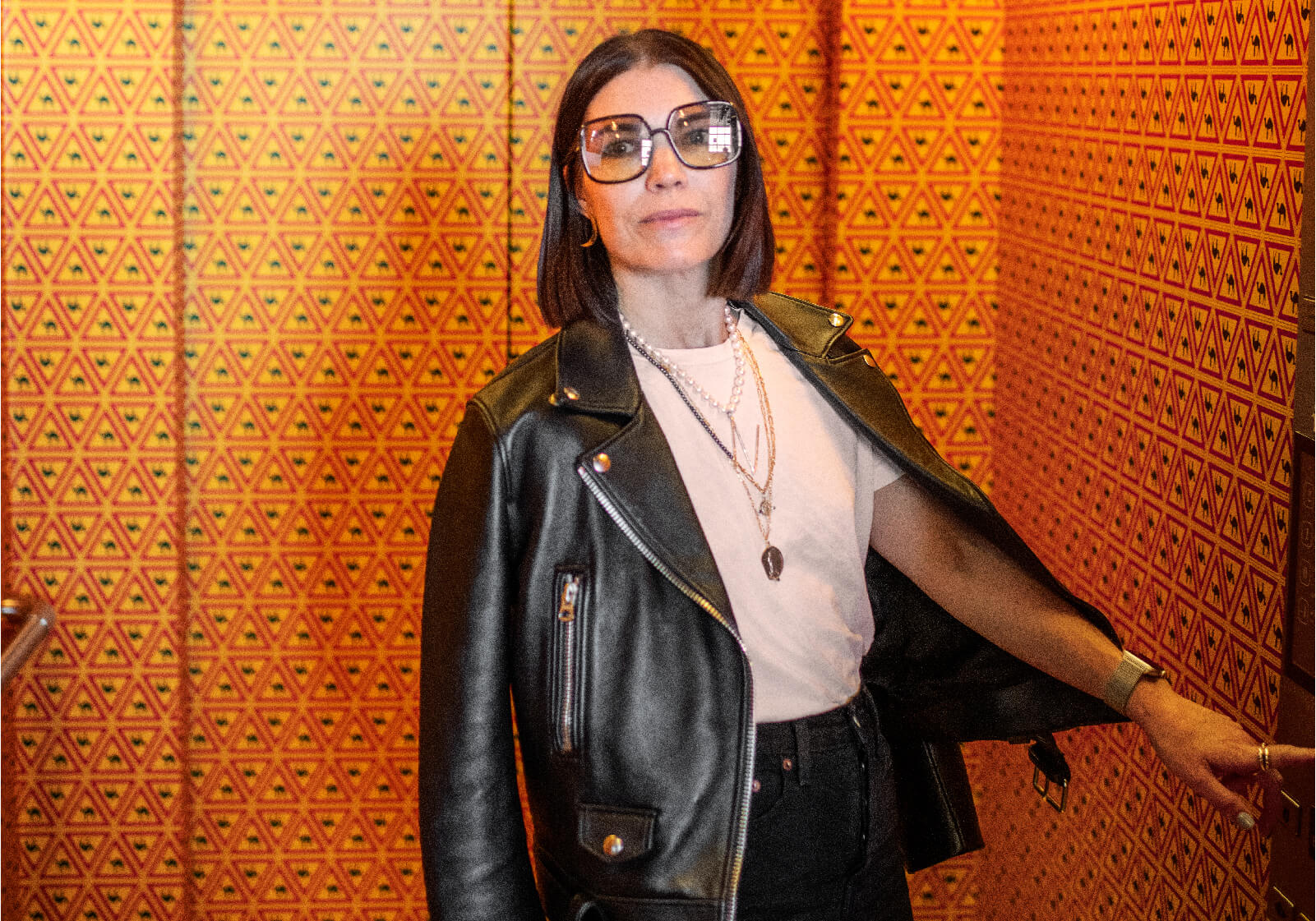Nina Lindvall står i en hiss med mönstrade tapeter iförd en skinnjacka och glasögon med stora bågar.