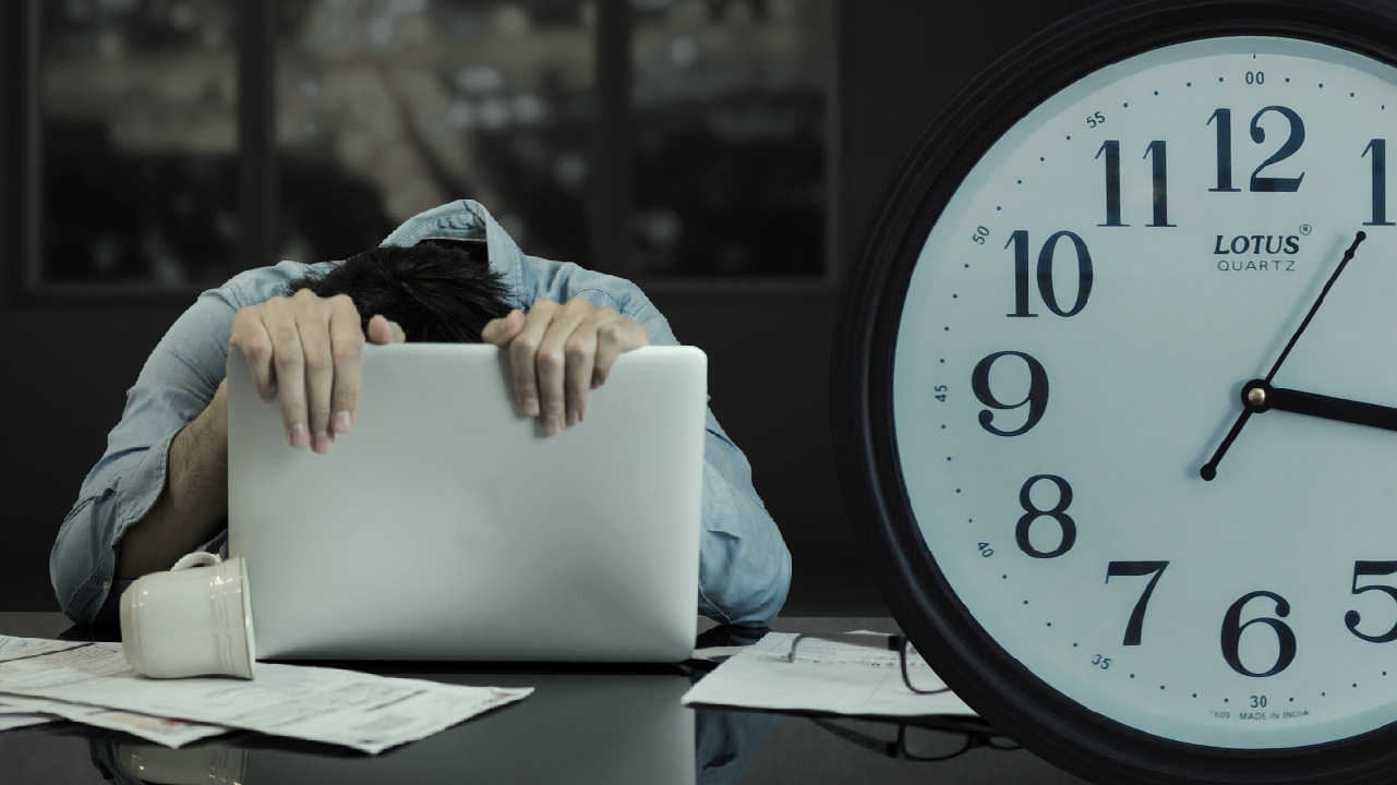 En person som ser mycket trött ut hänger över en dator. En klocka syns till höger i bilden.