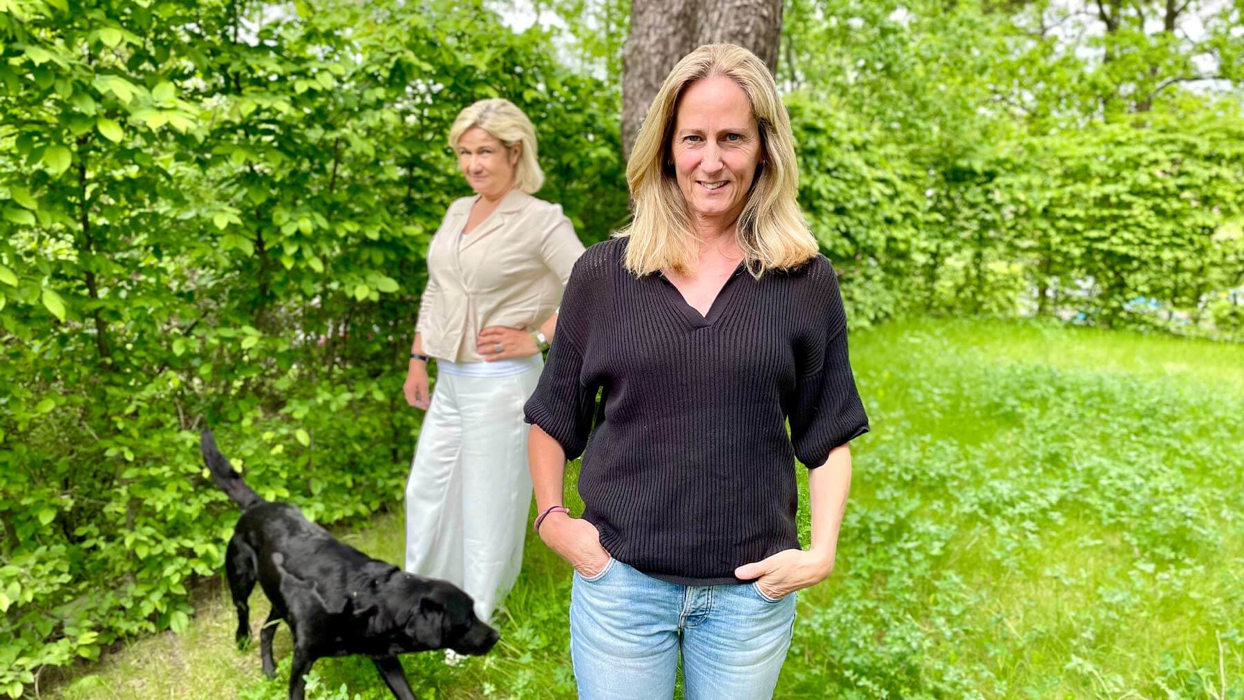 Katarina Ageborg i förgrunden och Cissi Elwin i bakgrunden. De står ute i en trädgård och vid Cissi står Katarinas svarta hund.