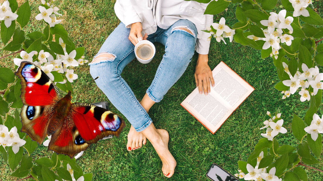 En somrig bild på en person som sitter i gräset (man ser bara personens ben) med en kopp kaffe i en handen och en den andra handen på en bok. Bredvid Till vänster i bilden syns en röd, vacker fjäril.