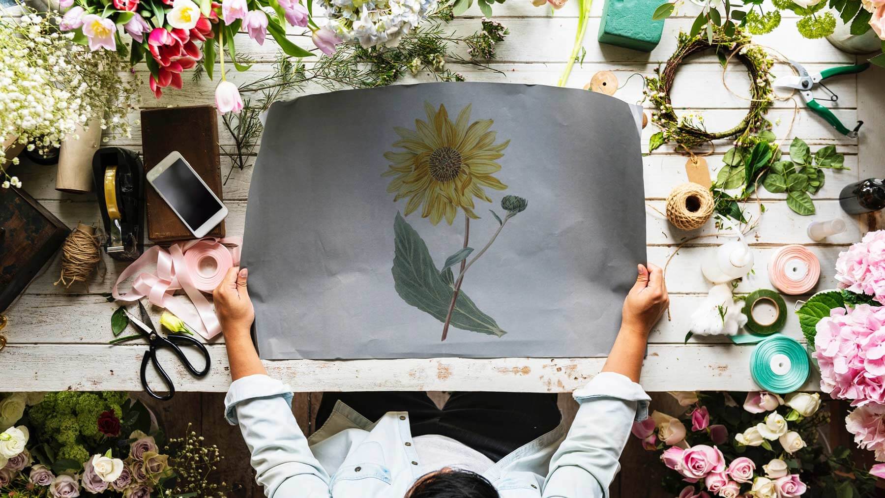 En kvinna håller upp en målad bild av en solros. Under bilden syns ett bord och vackra blommor.