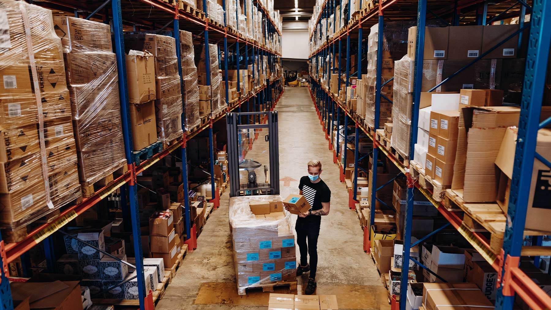 En ung kille med munskydd arbetar ensam i en stor lagerlokal.