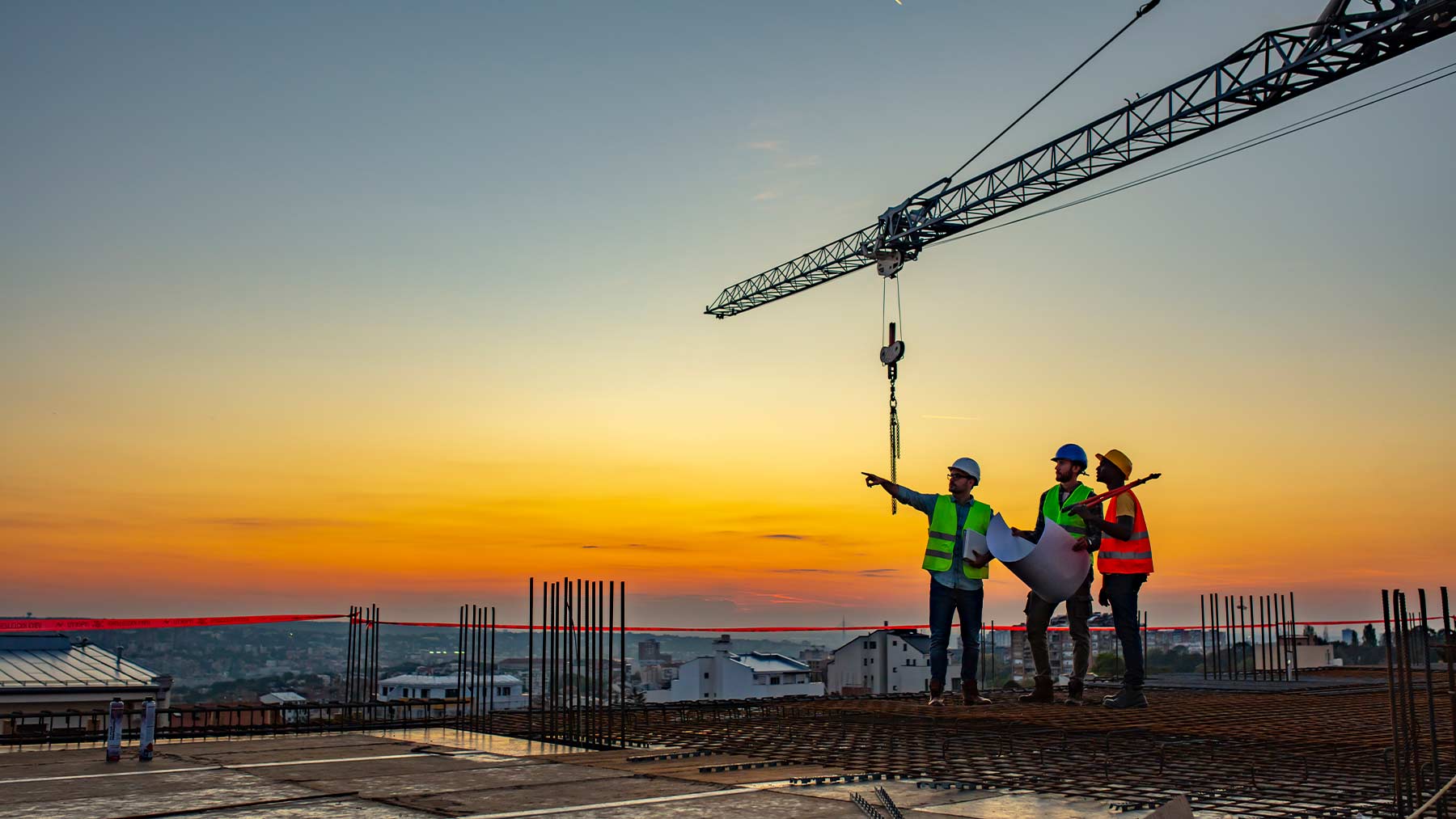 Tre män som arbetar på en byggarbetsplats står och pratar. Det är solnedgång.