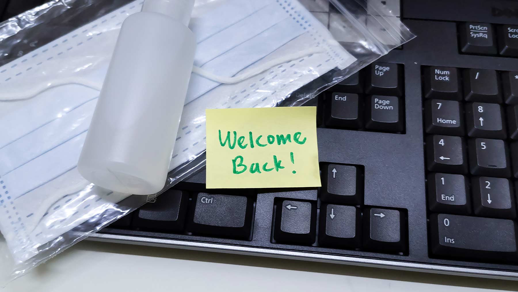 Ett tangentbord och ovanpå ligger ett munskydd och en liten flaska handsprit. Och en lapp med texten: "Välkommen tillbaka!".