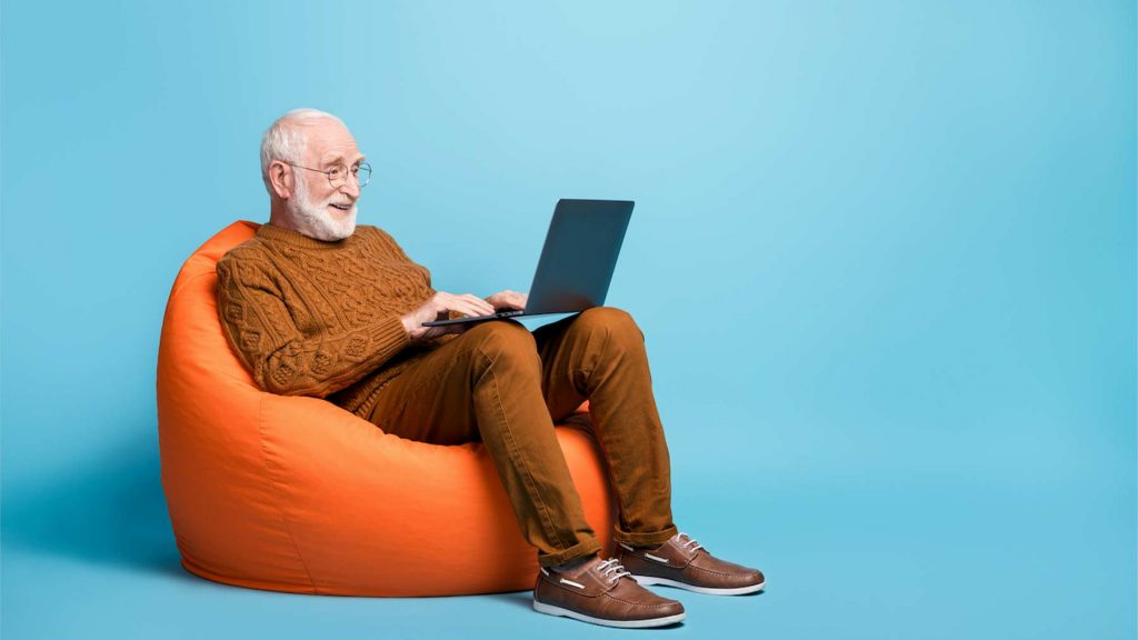 En äldre man sitter i en fåtölj med en bärbar dator i knäet.
