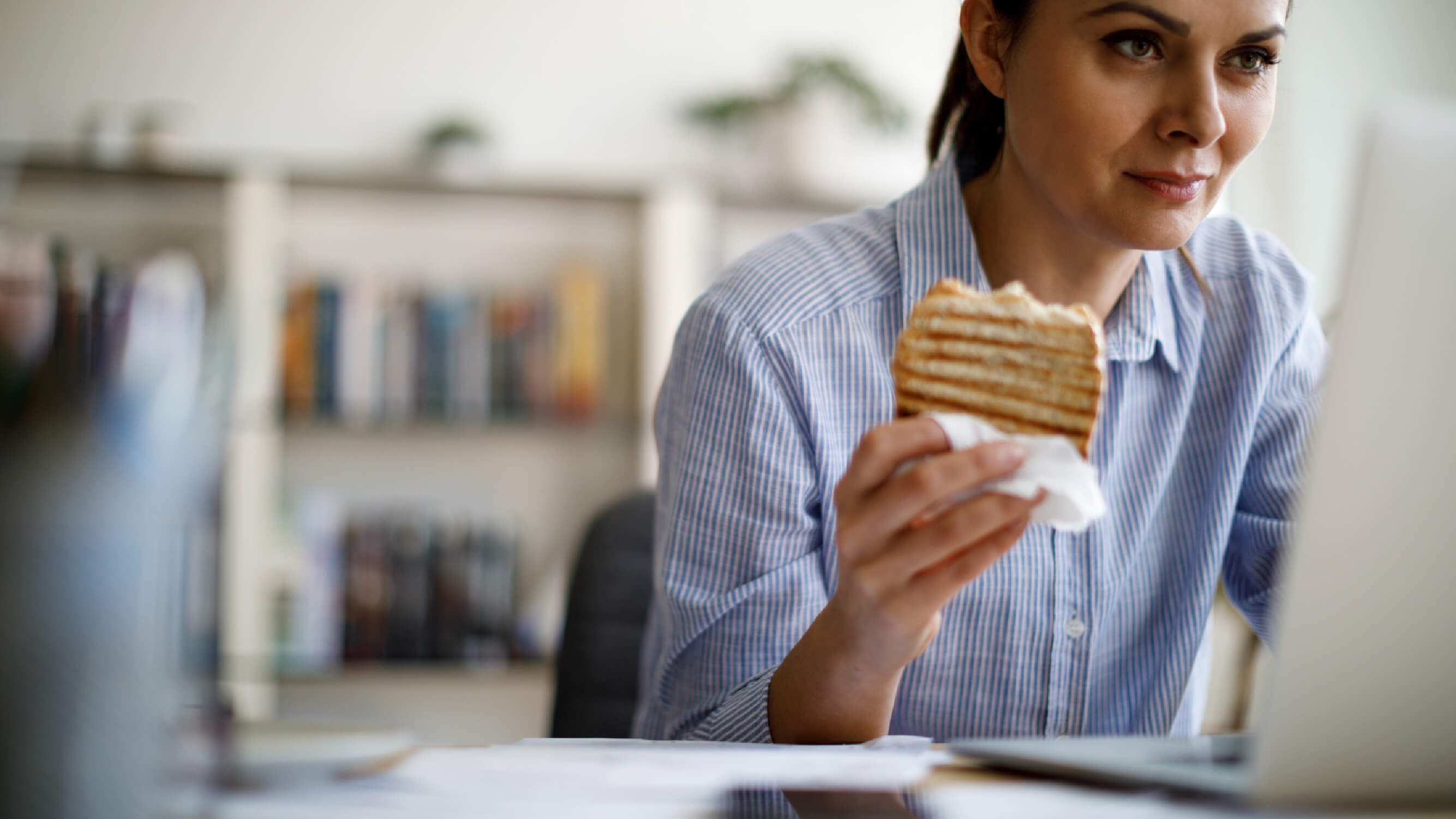 En ung kvinna sitter framför sin dator och äter lunch, i form av en smörgås.