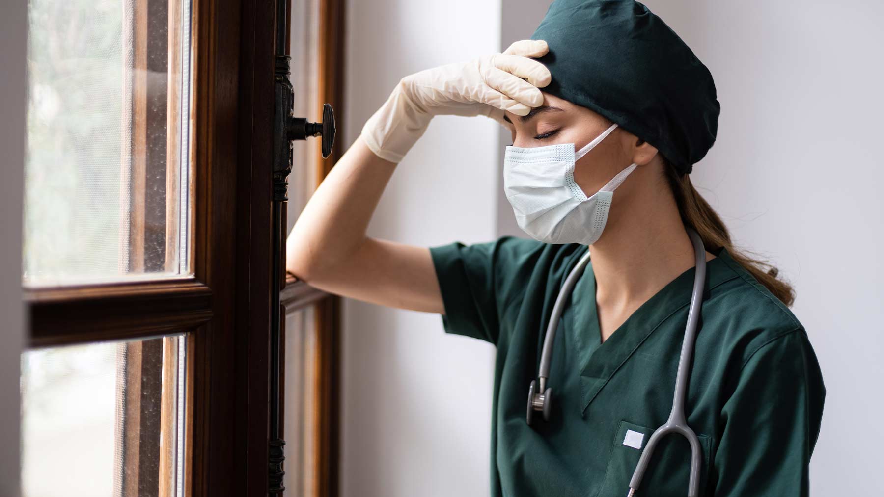 En kvinnlig läkare iförd munskydd tittar ut genom ett fönster och ser stressad ut.